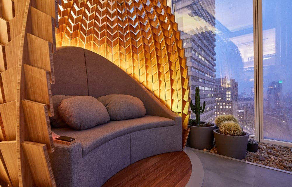 A cushioned reading nook inside a bamboo pod, lit amber, with the city through the window beside it