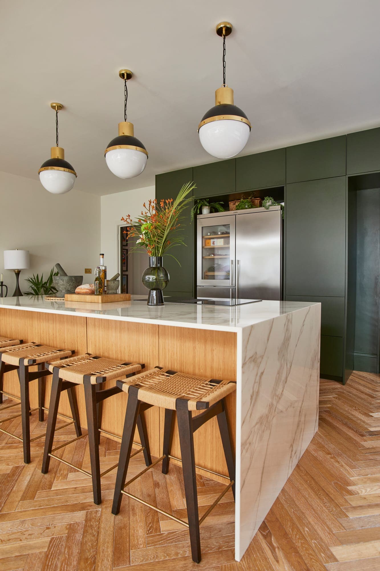 Kitchen island with bamboo fronts and a marble waterfall top, woven stools and globe pendants above