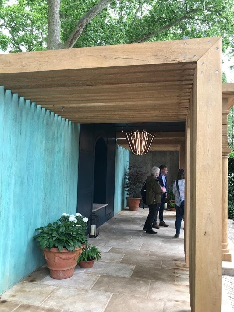 The verdigris wall running alongside an oak-framed pavilion, with potted white flowers at its foot