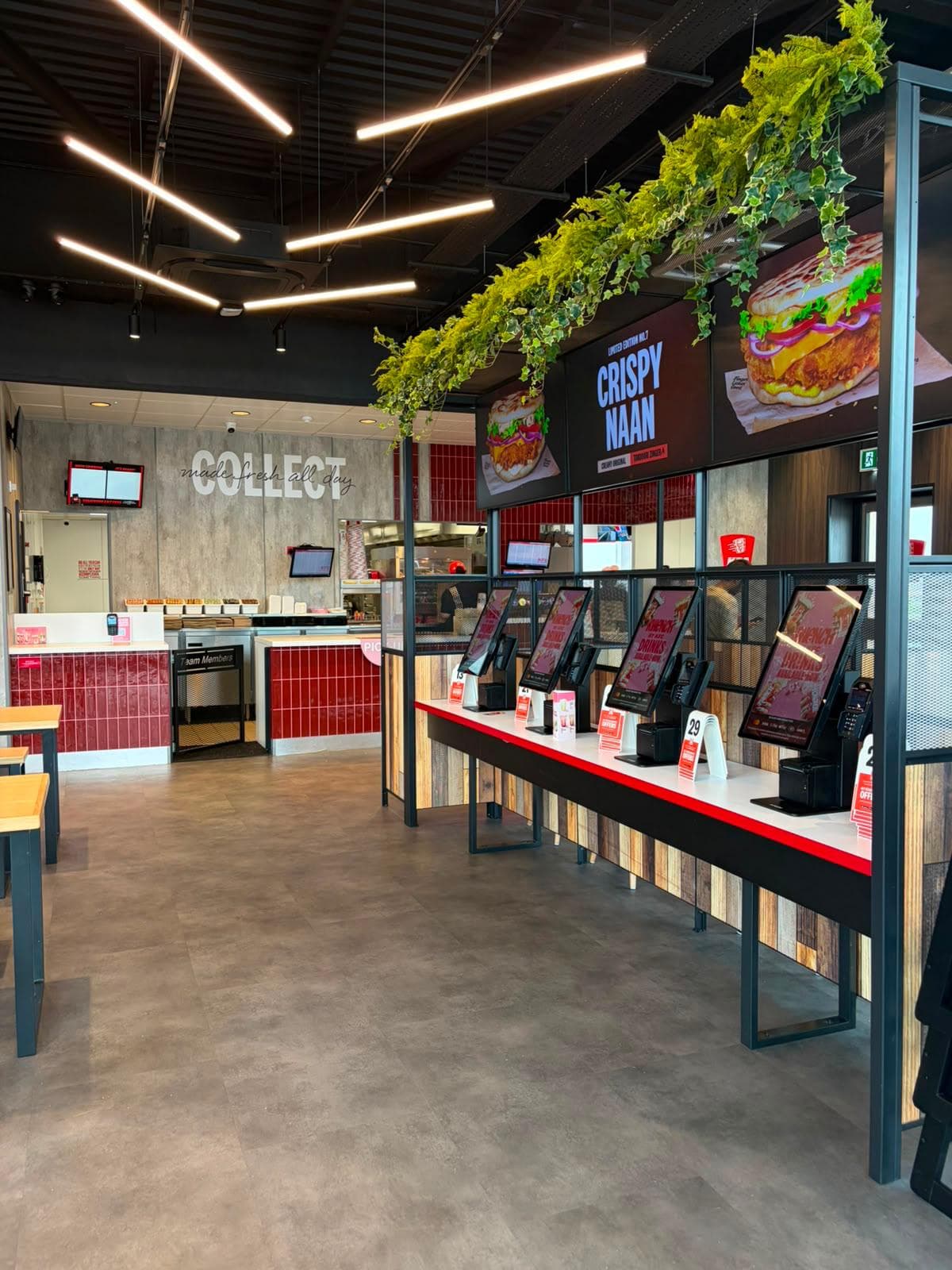 Self-order kiosks on a timber-clad counter under hanging greenery, with the tiled collect point beyond