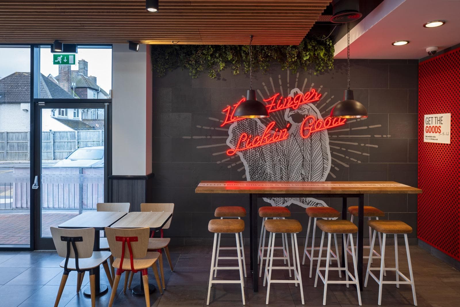 Timber high table and stools beneath a neon 'Finger Lickin' Good' sign, with dining tables by the window